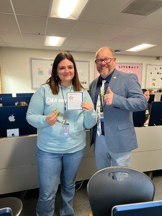 Woman in blue sweatshirt and man in blazer pose together, holding a certificate. Computer lab.