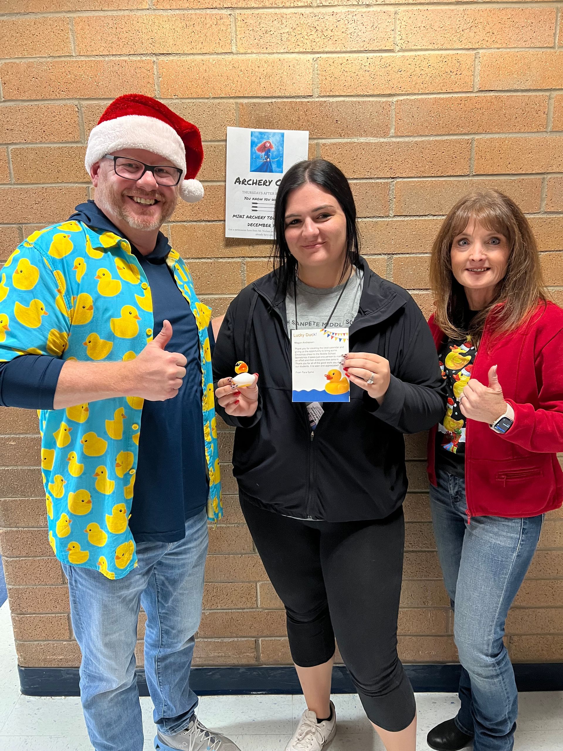 A man in a santa hat is standing next to two women holding cupcakes and giving a thumbs up.