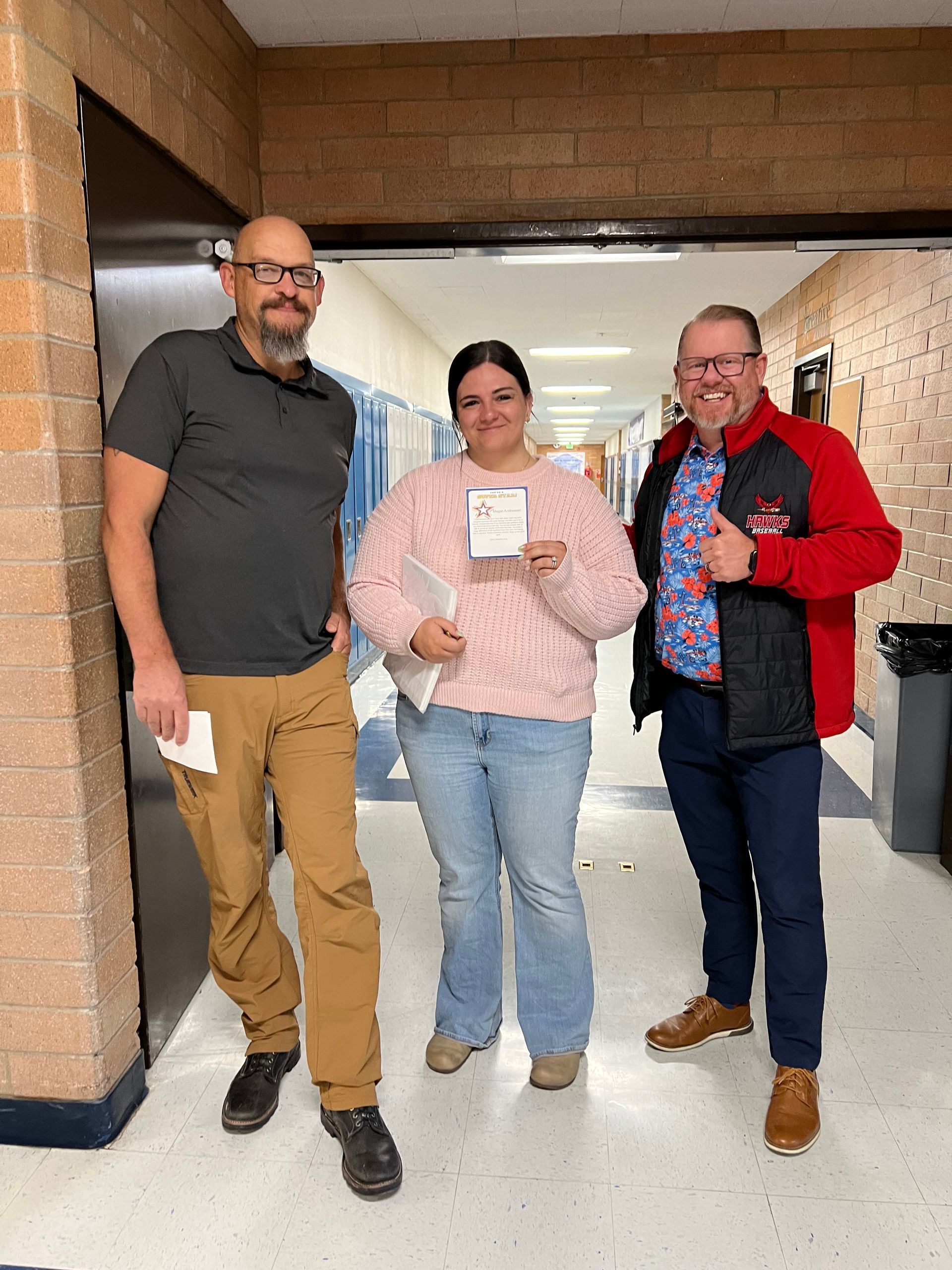 Three people standing in a hallway, smiling and holding small papers, with brick walls and blue doors nearby.