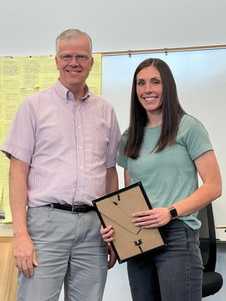 Two people smiling and posing indoors, holding a framed certificate in front of a whiteboard.