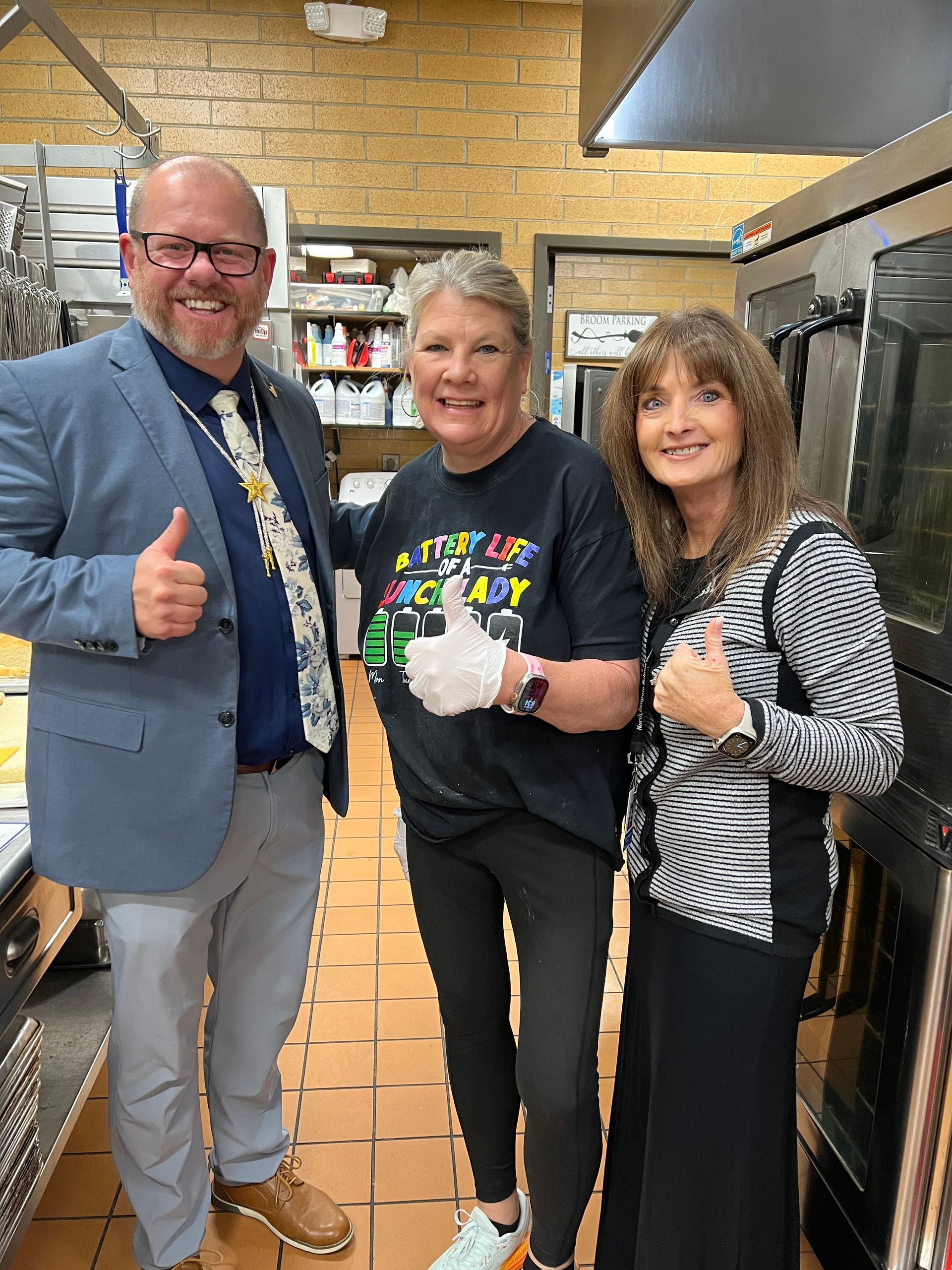Three people in a kitchen give thumbs up. One wears a blue blazer. Another wears a black shirt, gloves, and black pants.