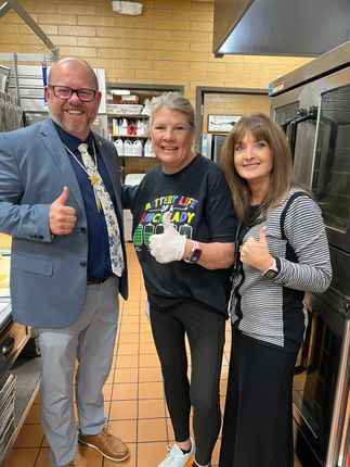 Three people in a kitchen give thumbs up. One wears a blazer, another gloves, and the third a striped shirt.