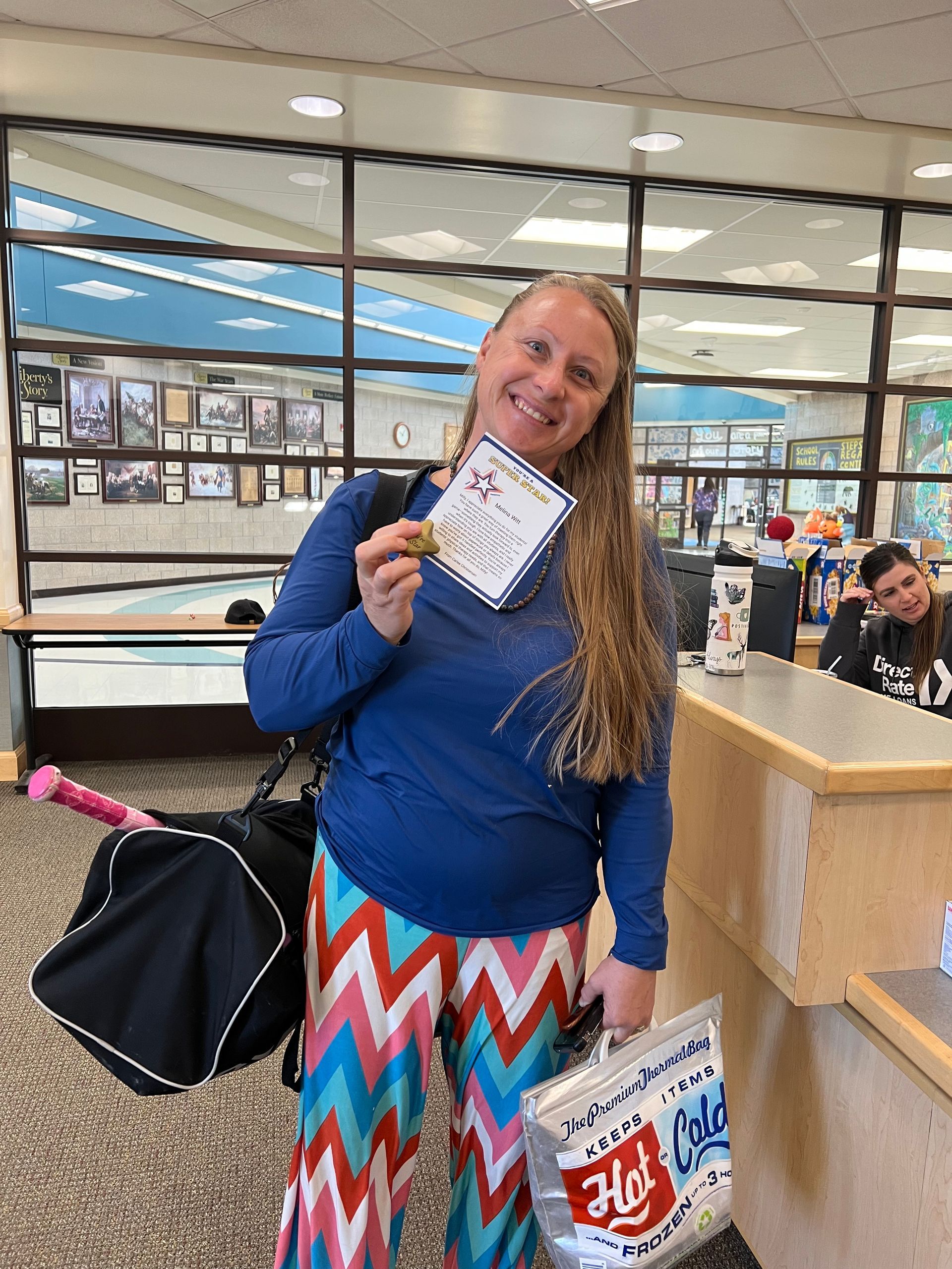 Woman holding a paper and bag with a smile, standing near a check-in desk, wearing blue and colorful pants.