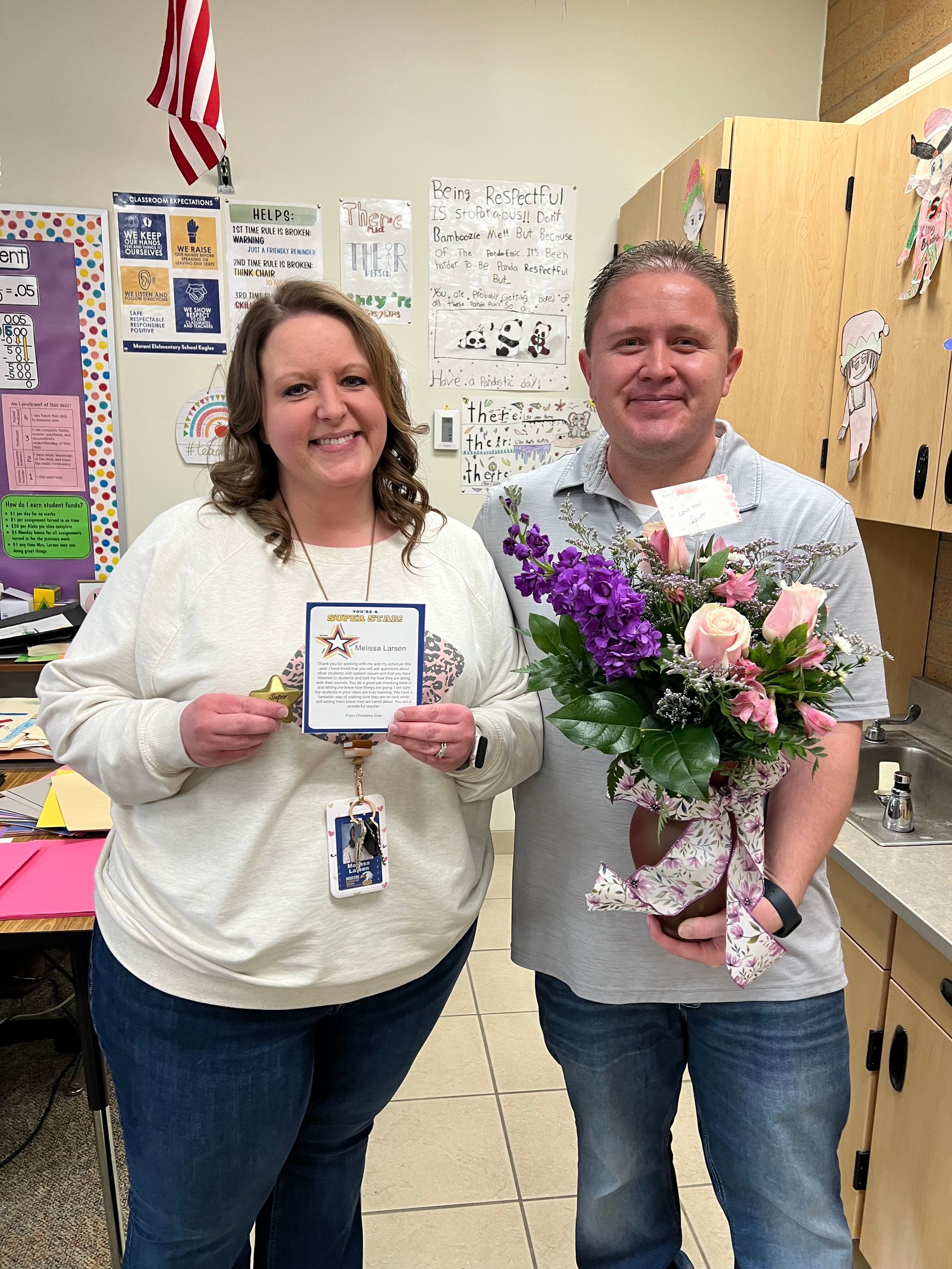 Two people holding flowers and a card, smiling in a classroom setting.
