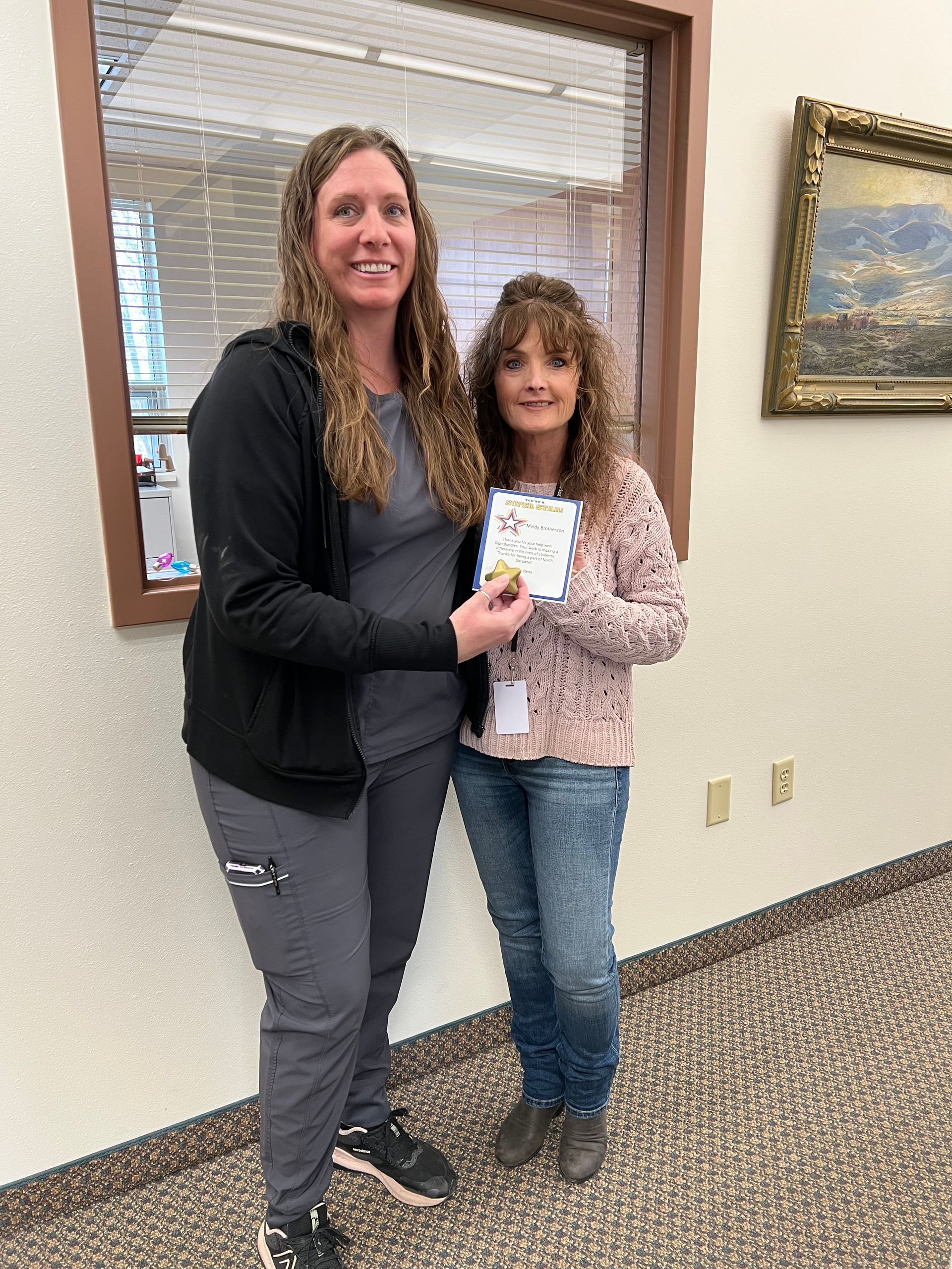 Two women, one in scrubs, holding a card. They smile inside a building.