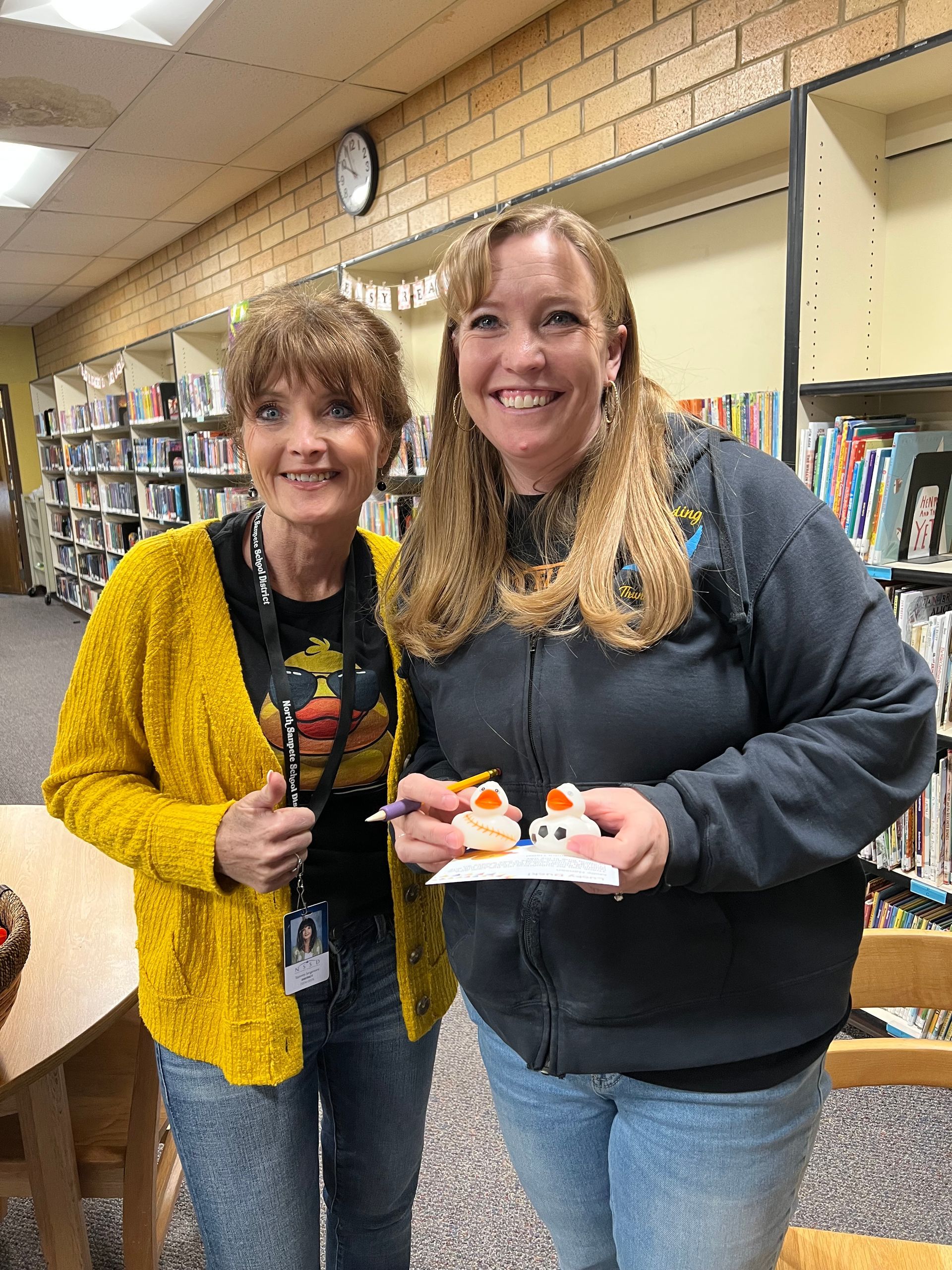 Two women are standing next to each other in a library holding cupcakes.