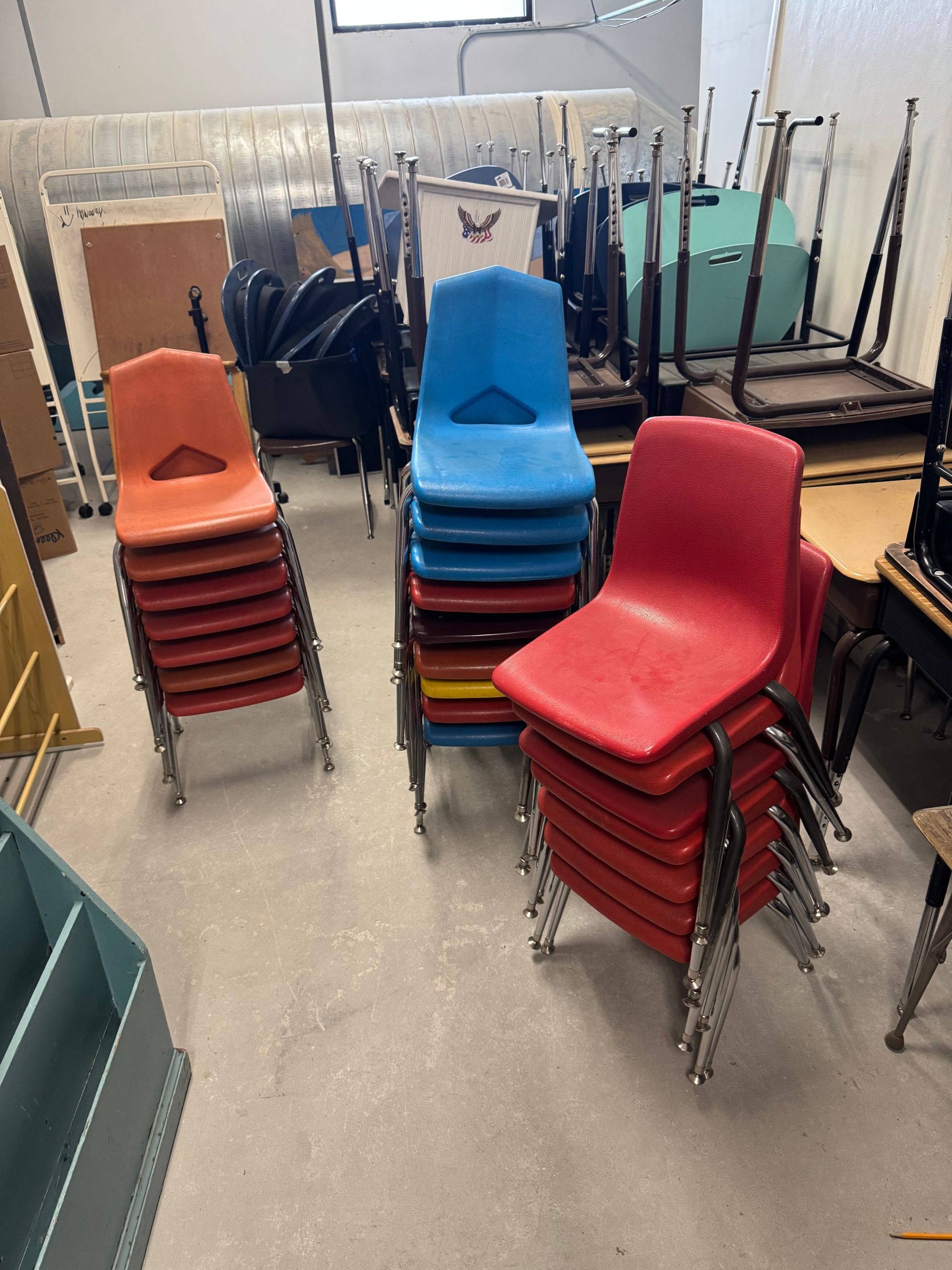 Stacks of colorful plastic chairs in a classroom storage area: orange, blue, and red.