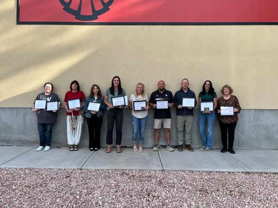 Nine people stand in a row holding certificates in front of a beige wall.