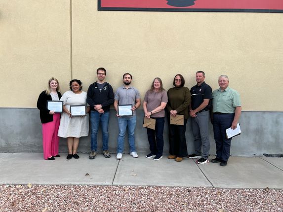 Eight people stand in a row outside a building, several holding certificates or awards.