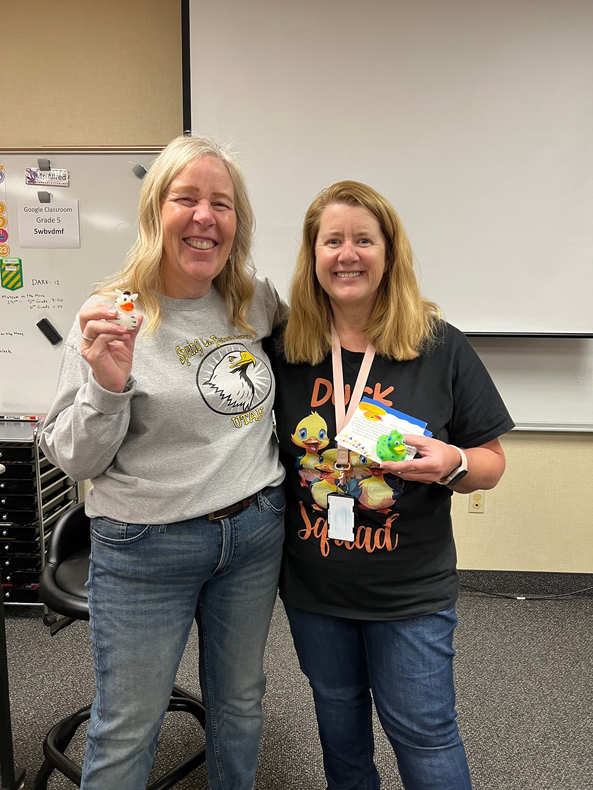 Two women are standing next to each other in a classroom and smiling.