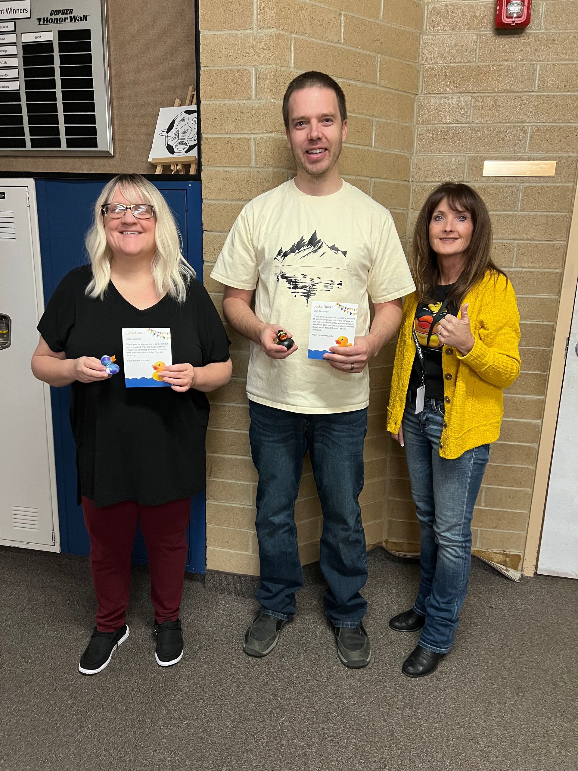 Three people are standing next to each other in a hallway holding cards.