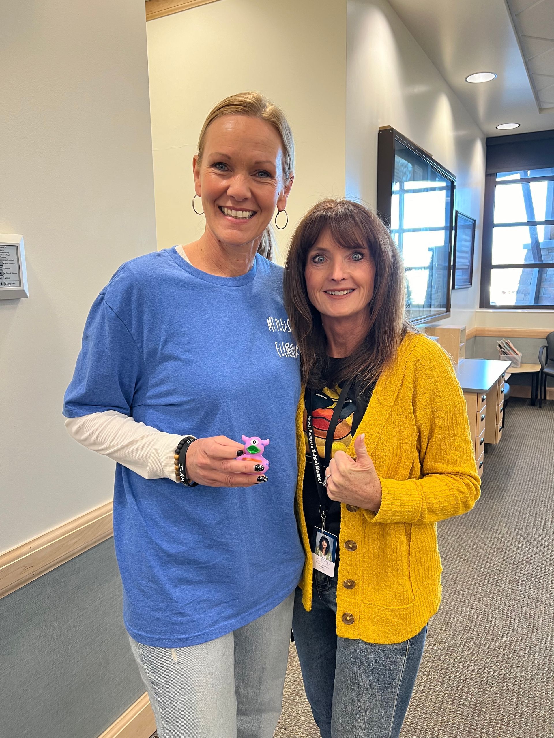 Two women are posing for a picture in a hallway . one of the women is wearing a blue shirt.