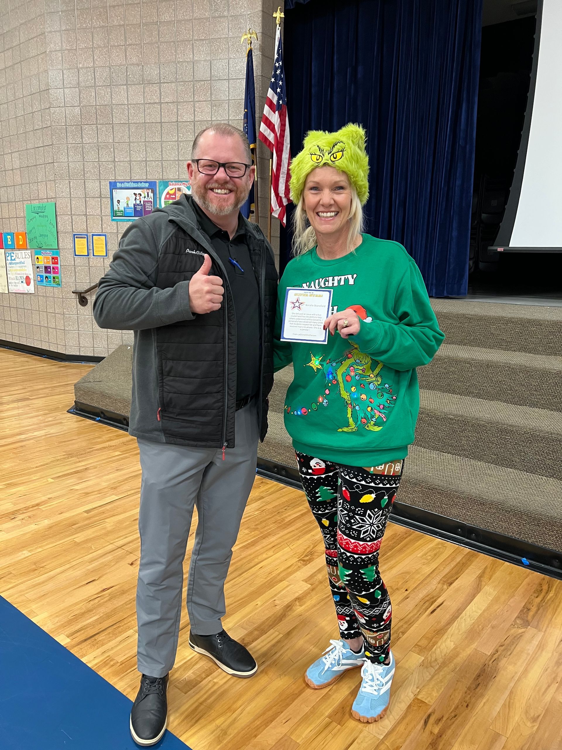 Man giving thumbs up stands with woman in a green sweater, holding a paper. Both are on a stage.