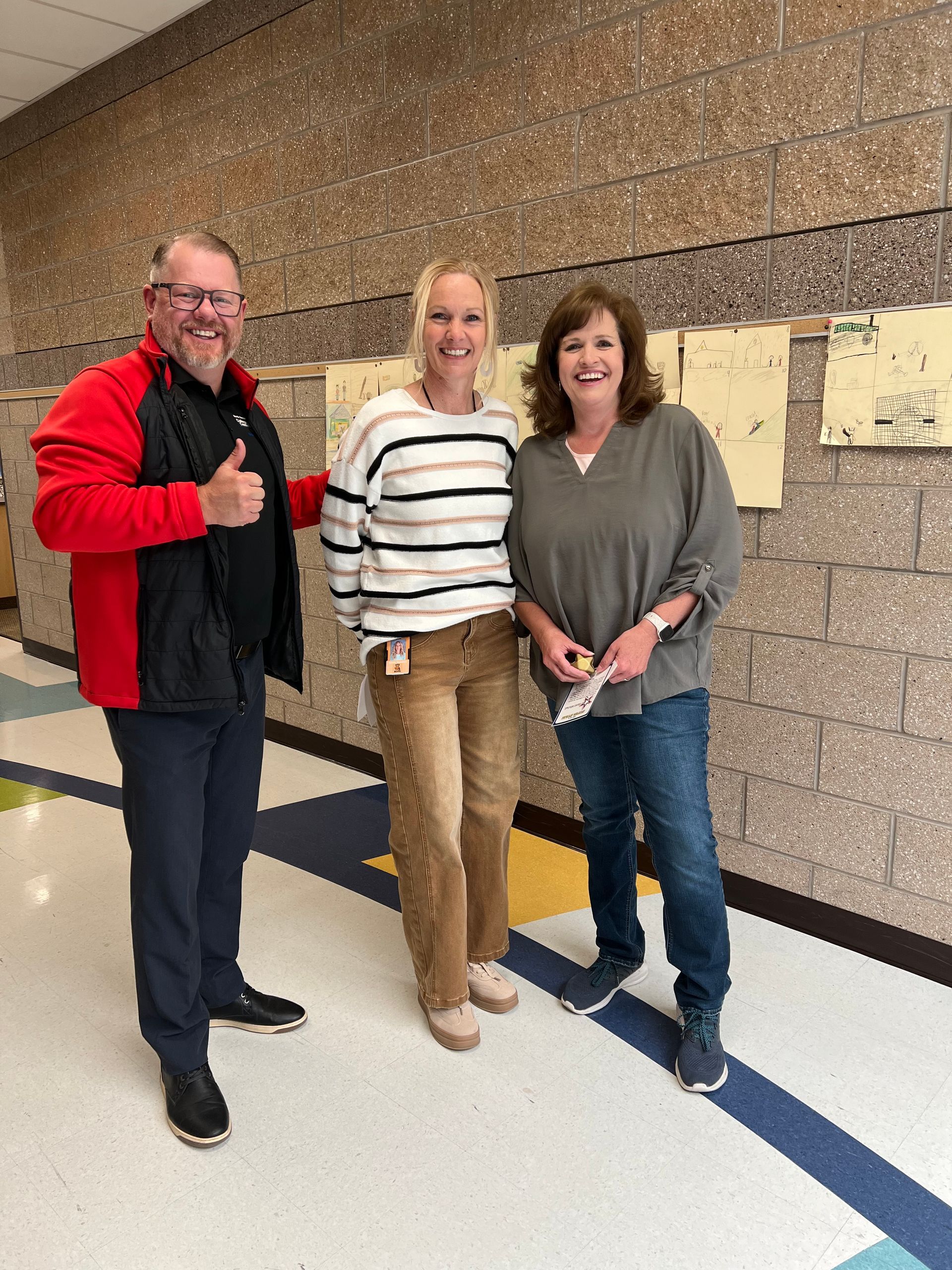 Three people standing in a hallway, one giving a thumbs-up and all smiling.