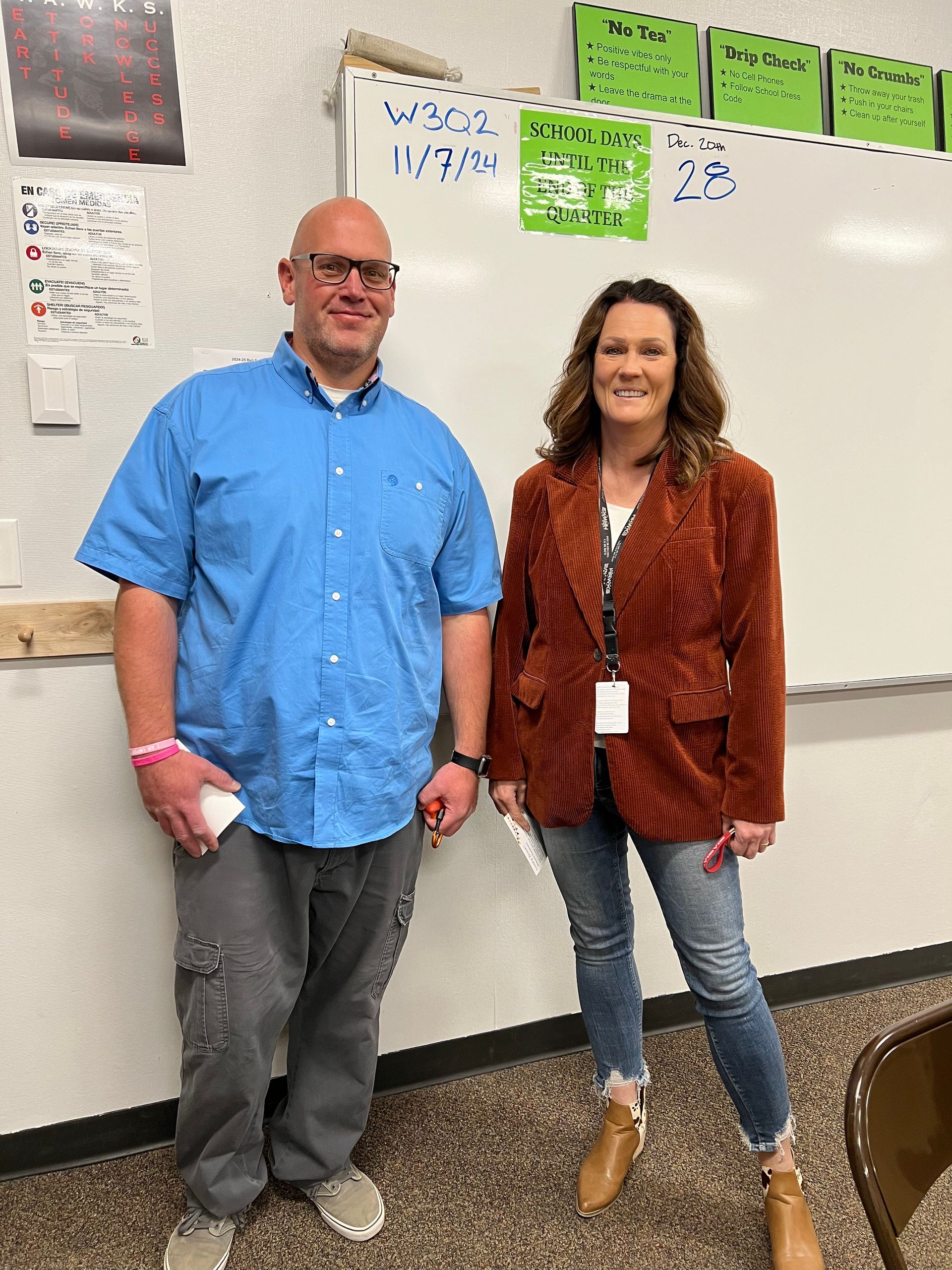 A man and a woman are standing next to each other in front of a whiteboard in a classroom.