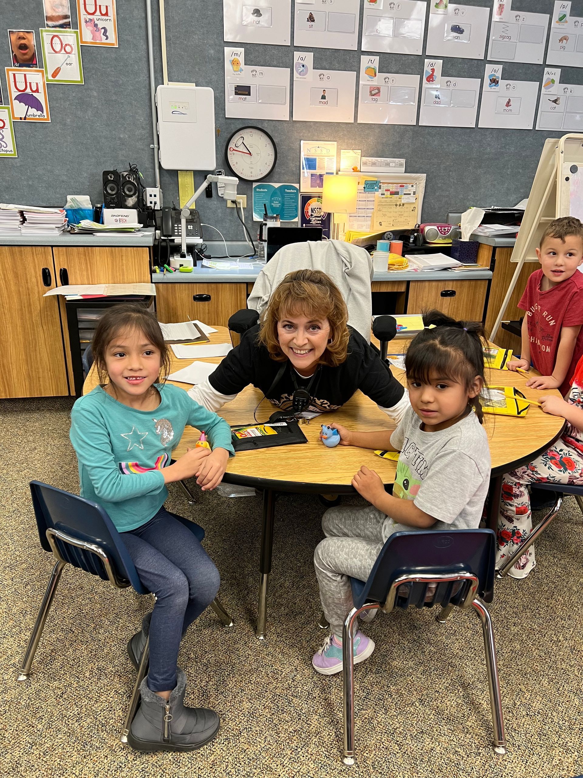 A woman is sitting at a table with a group of children in a classroom.