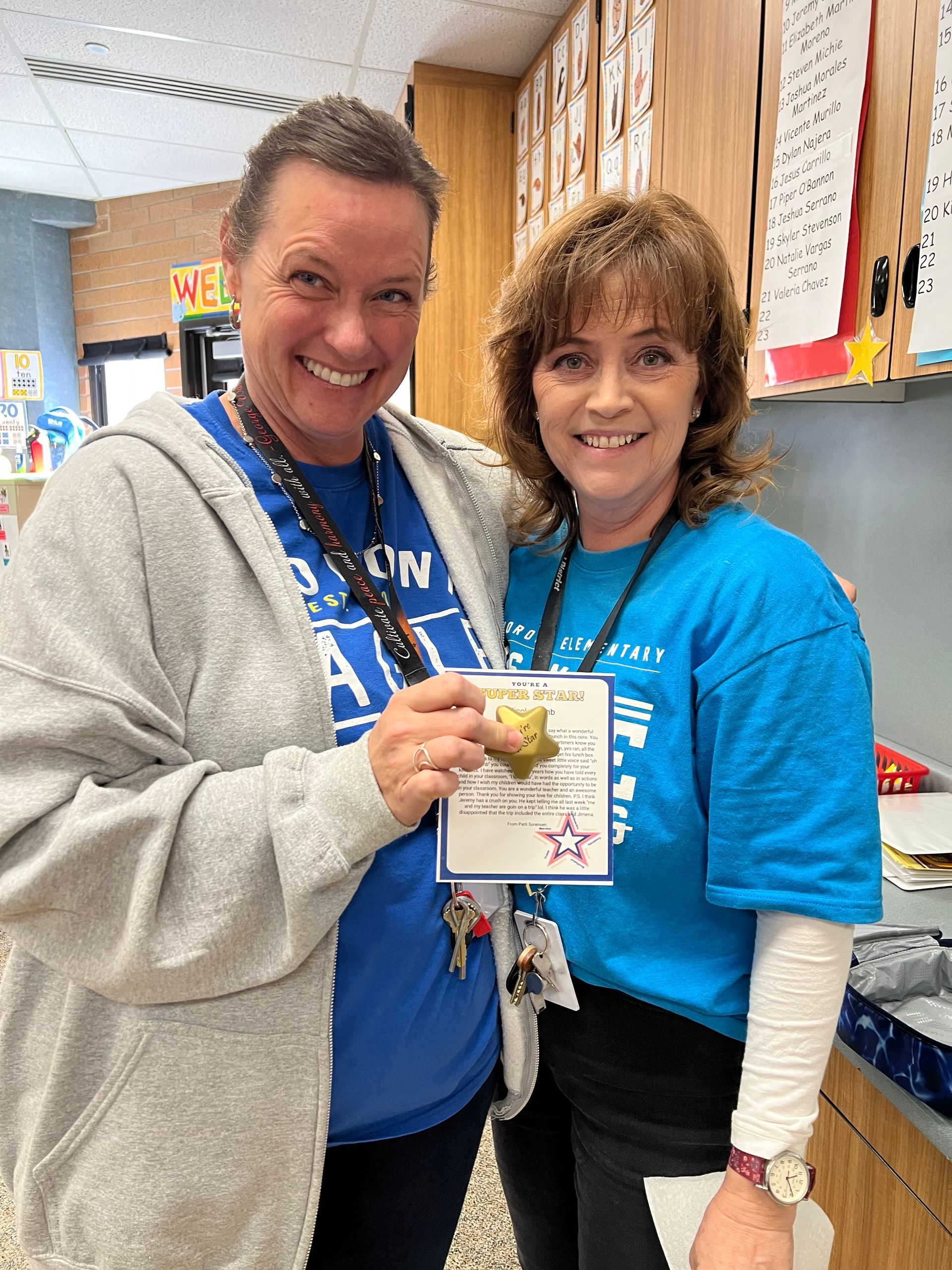 Two women smiling, holding a star-shaped award in a classroom. One wears a blue shirt, the other a grey hoodie.