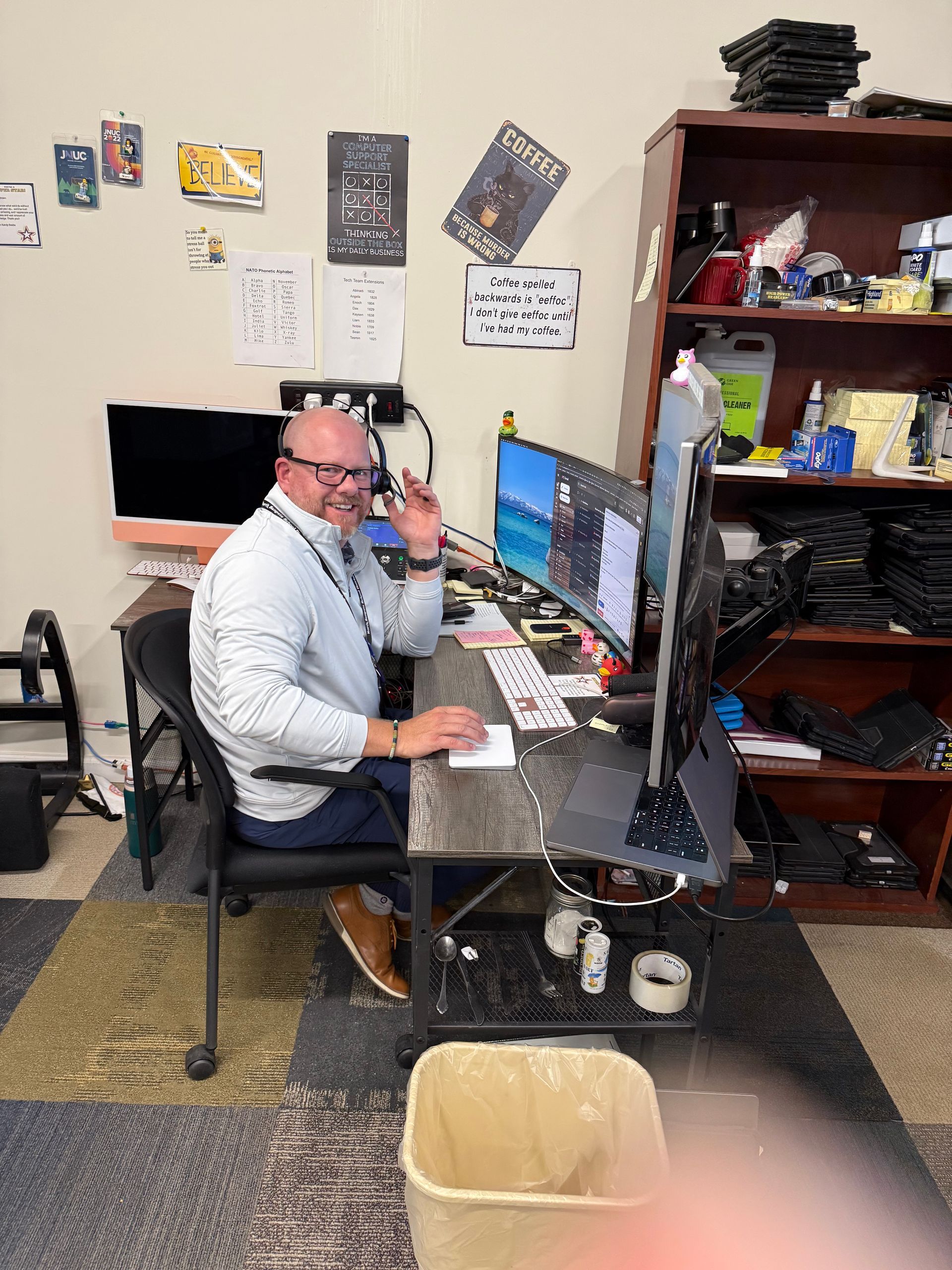 Man at desk, waving. Computer monitors, office setting.
