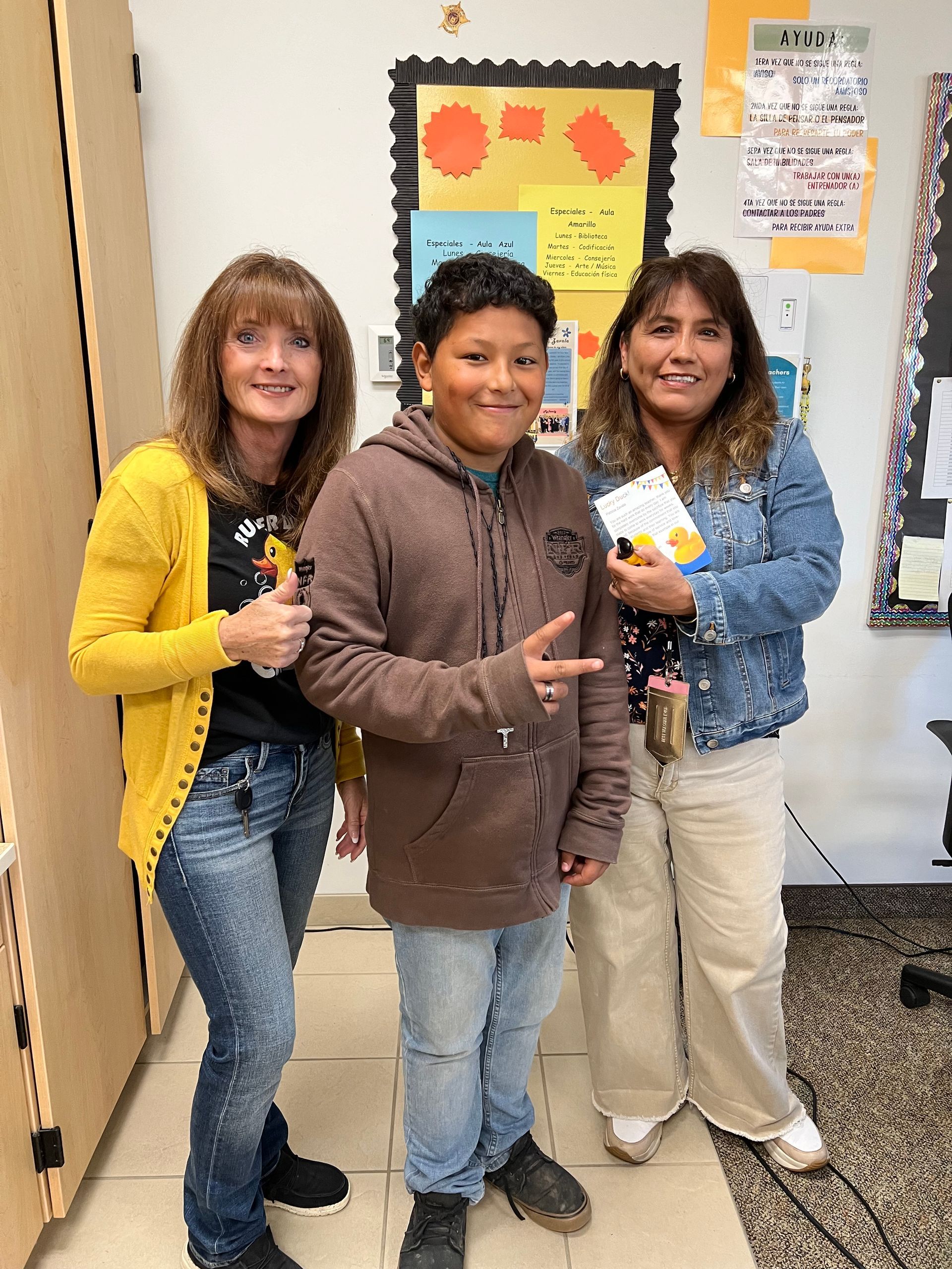 Two women and a boy are standing next to each other in front of a bulletin board.