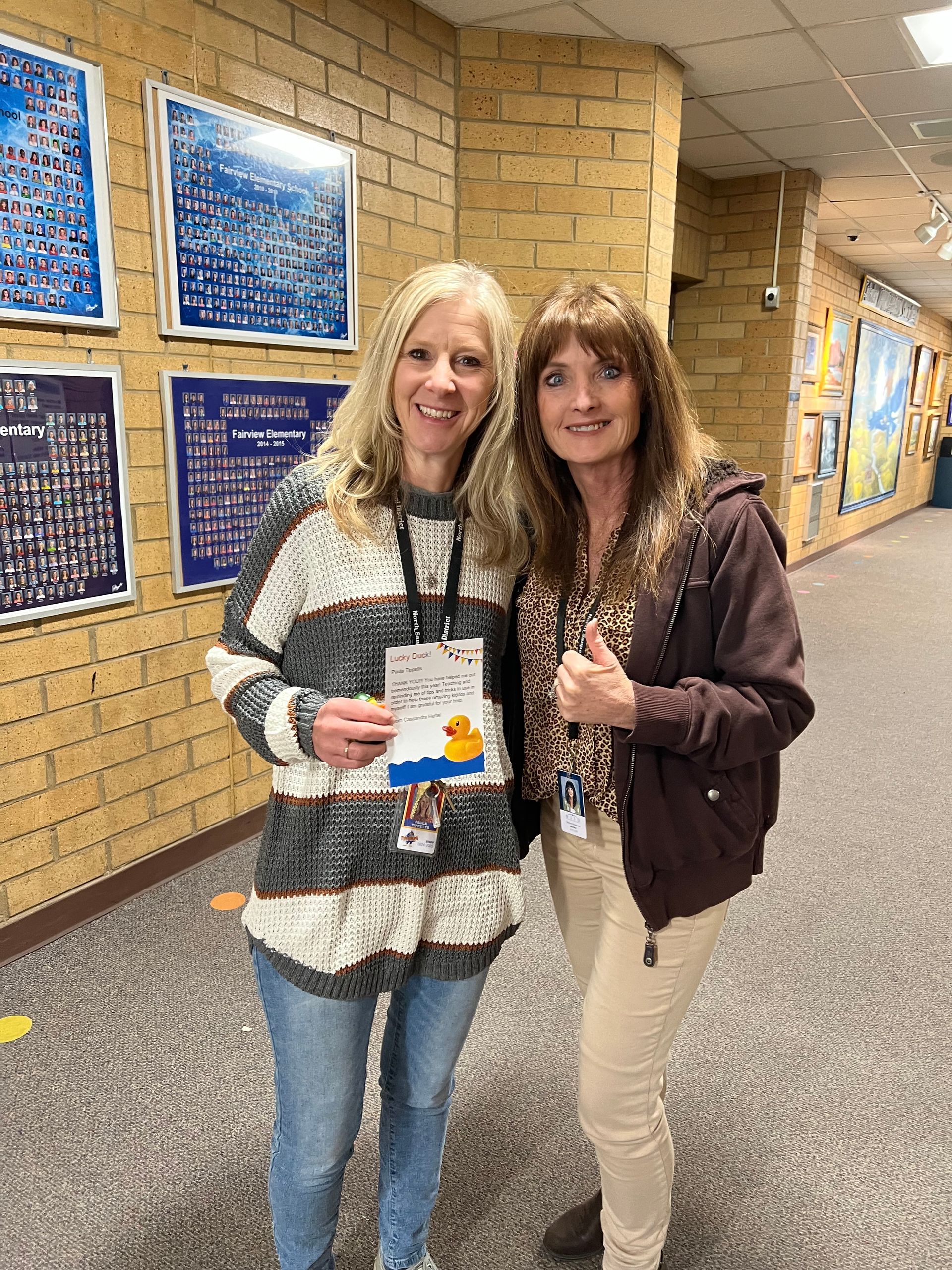 Two women are standing next to each other in a hallway giving a thumbs up.