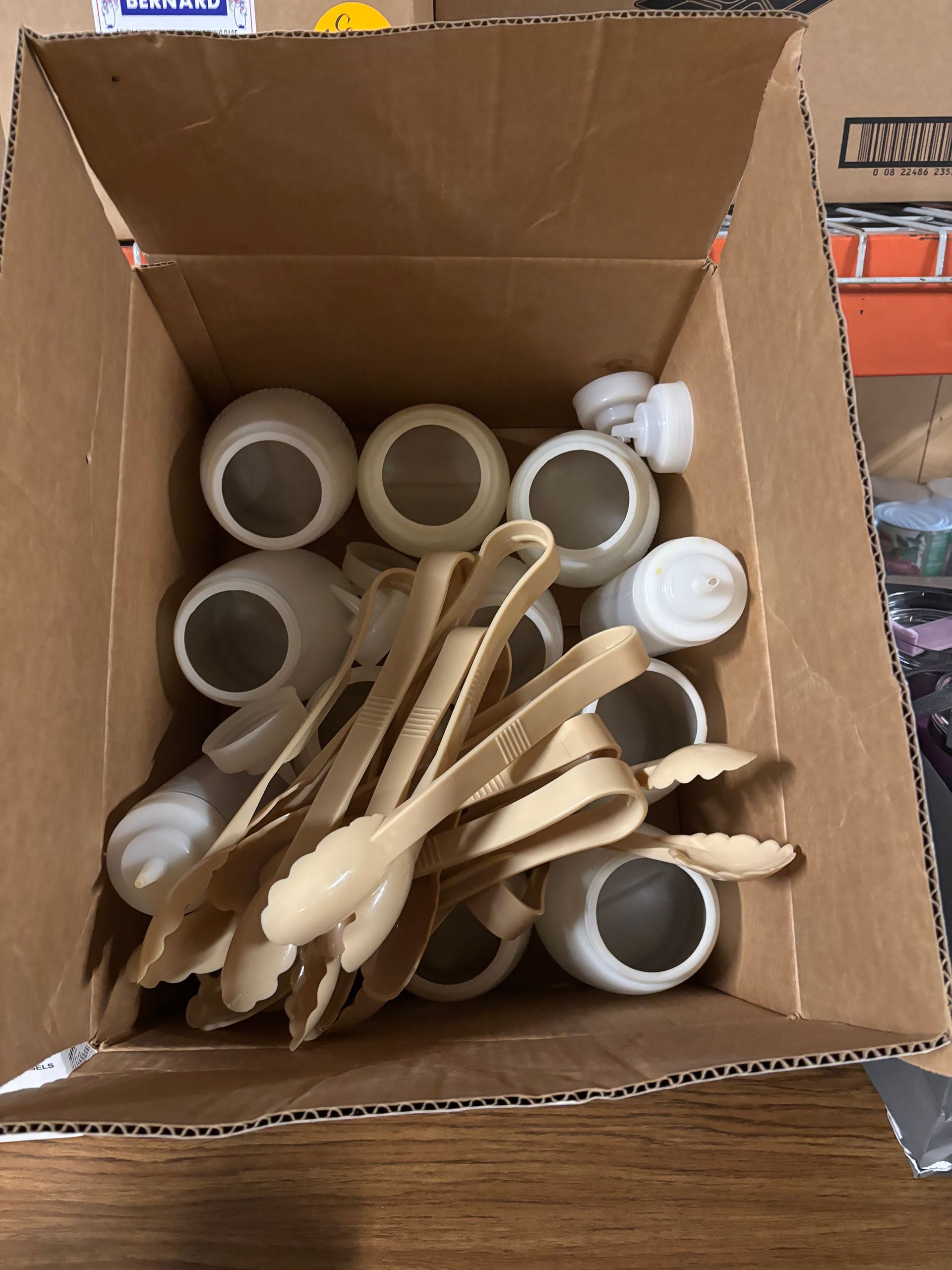 A cardboard box containing several white plastic squeeze bottles and a set of beige plastic serving tongs.
