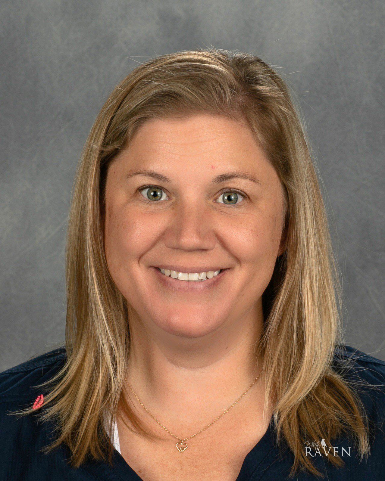 A person with shoulder-length blonde hair, wearing a dark blue top and a delicate gold necklace, smiles against a gray.