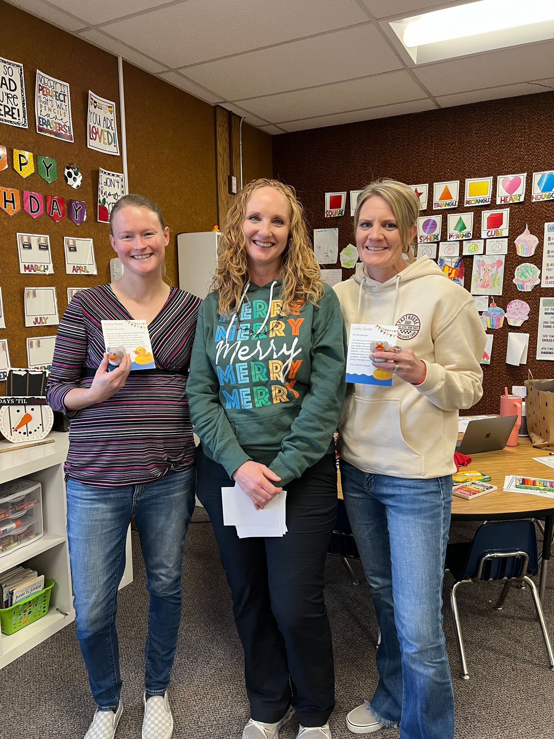 Three women are standing next to each other in a classroom holding papers.