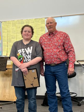 Two people standing in a classroom, one holding a plaque, with a whiteboard behind them.