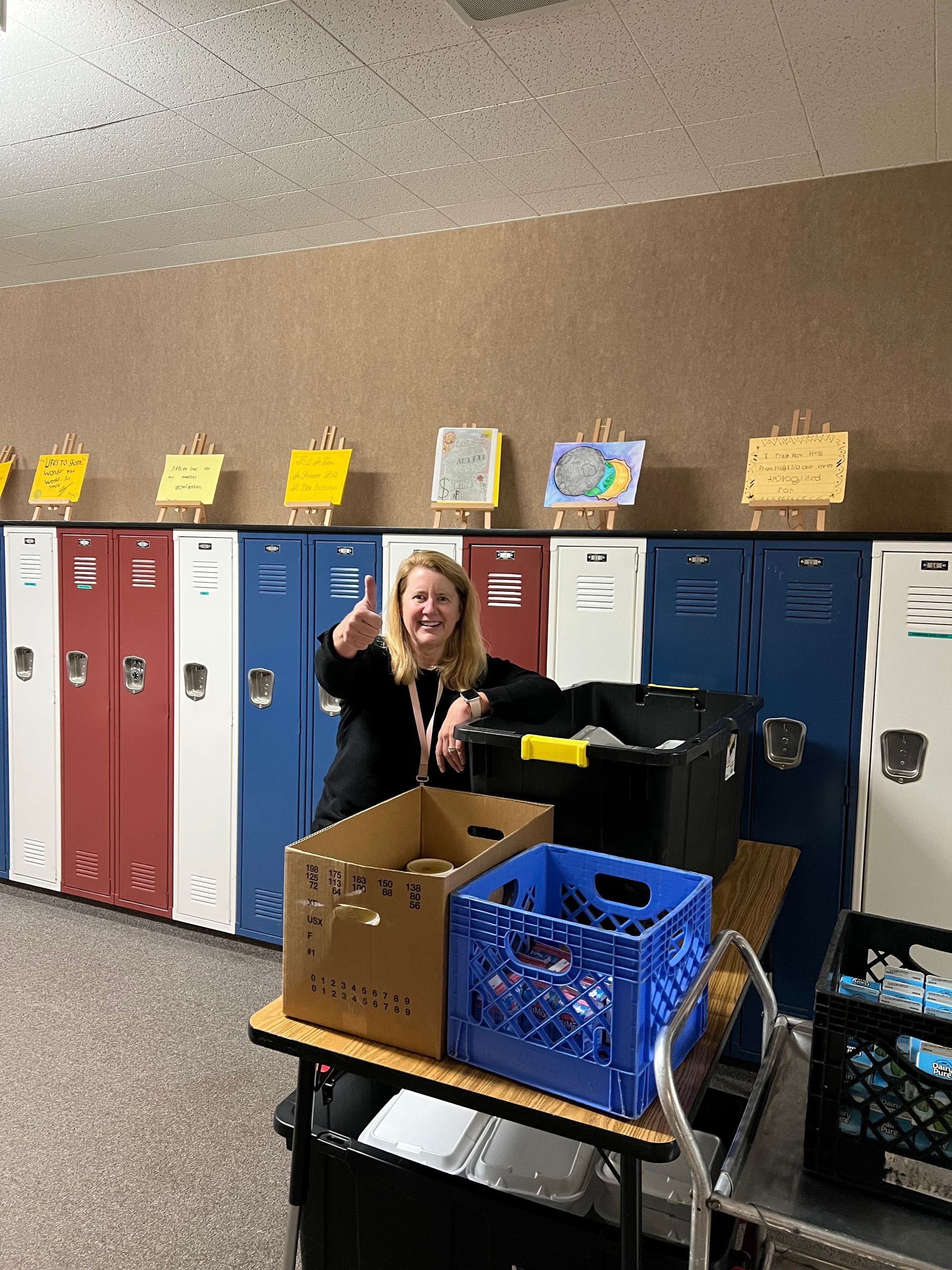 Woman giving thumbs up stands by a cart with crates near lockers with displayed artwork.