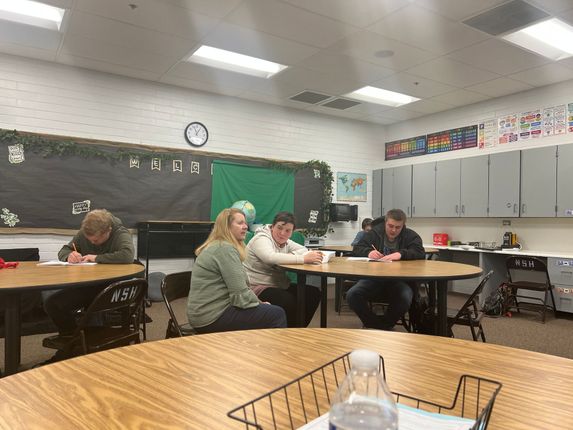 People seated at tables in a classroom, writing. Decorations on the wall, including a clock and artwork.