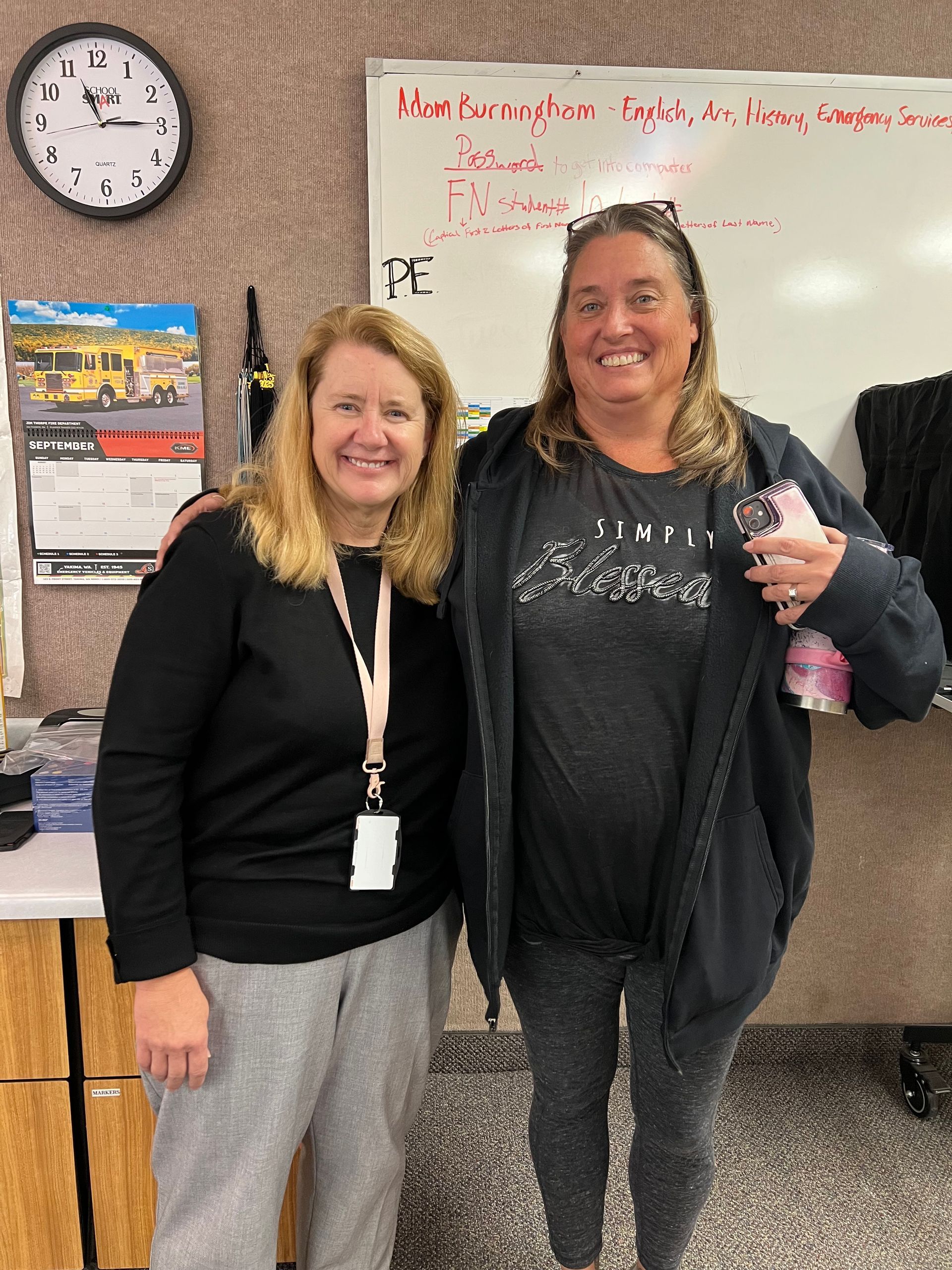 Two women smile, standing together indoors. One wears a black sweater, the other a dark hoodie and patterned leggings. A clock and whiteboard are in the background.