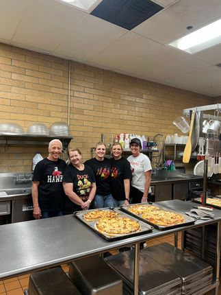 Five people in matching shirts stand behind a stainless steel table in a commercial kitchen with two trays of pizza.