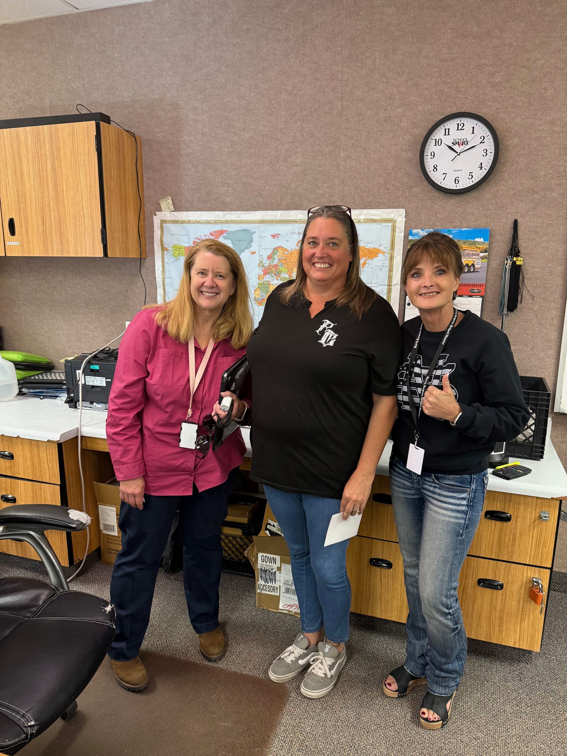 Three women smiling in a classroom. One in pink, one in black, and one in black and jeans. A world map is behind them.