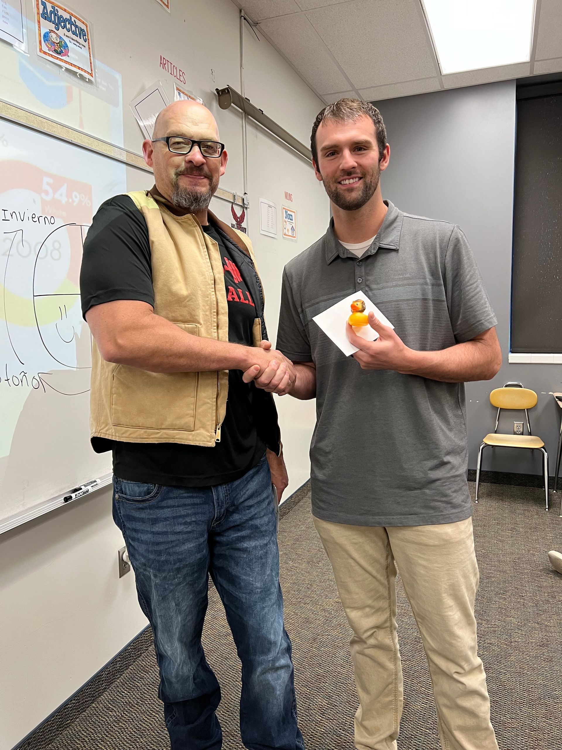 Two men are shaking hands in a classroom in front of a whiteboard.