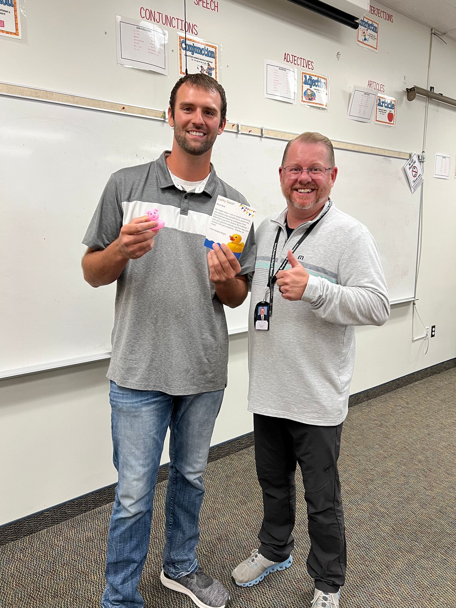 Two men are standing next to each other in a classroom holding papers.