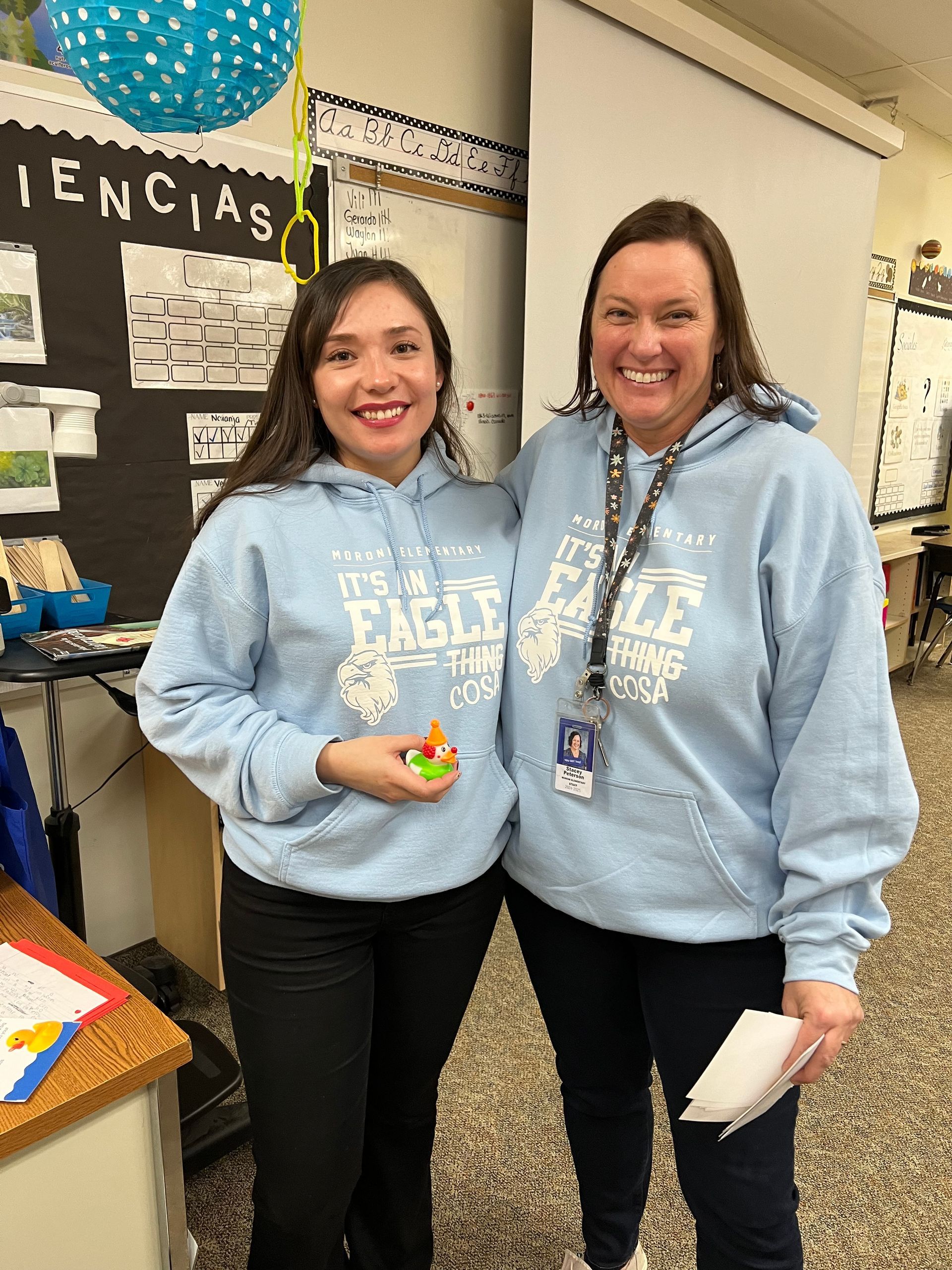 Two women wearing blue sweatshirts are standing next to each other in a classroom.