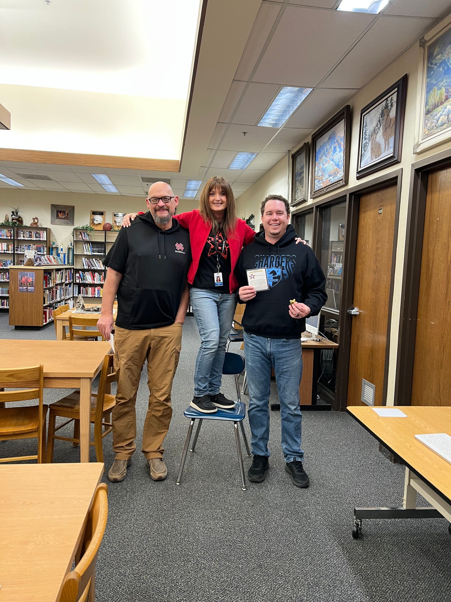 Three people in a library, one standing on a chair. Smiling, holding items. Wooden tables and bookshelves visible.