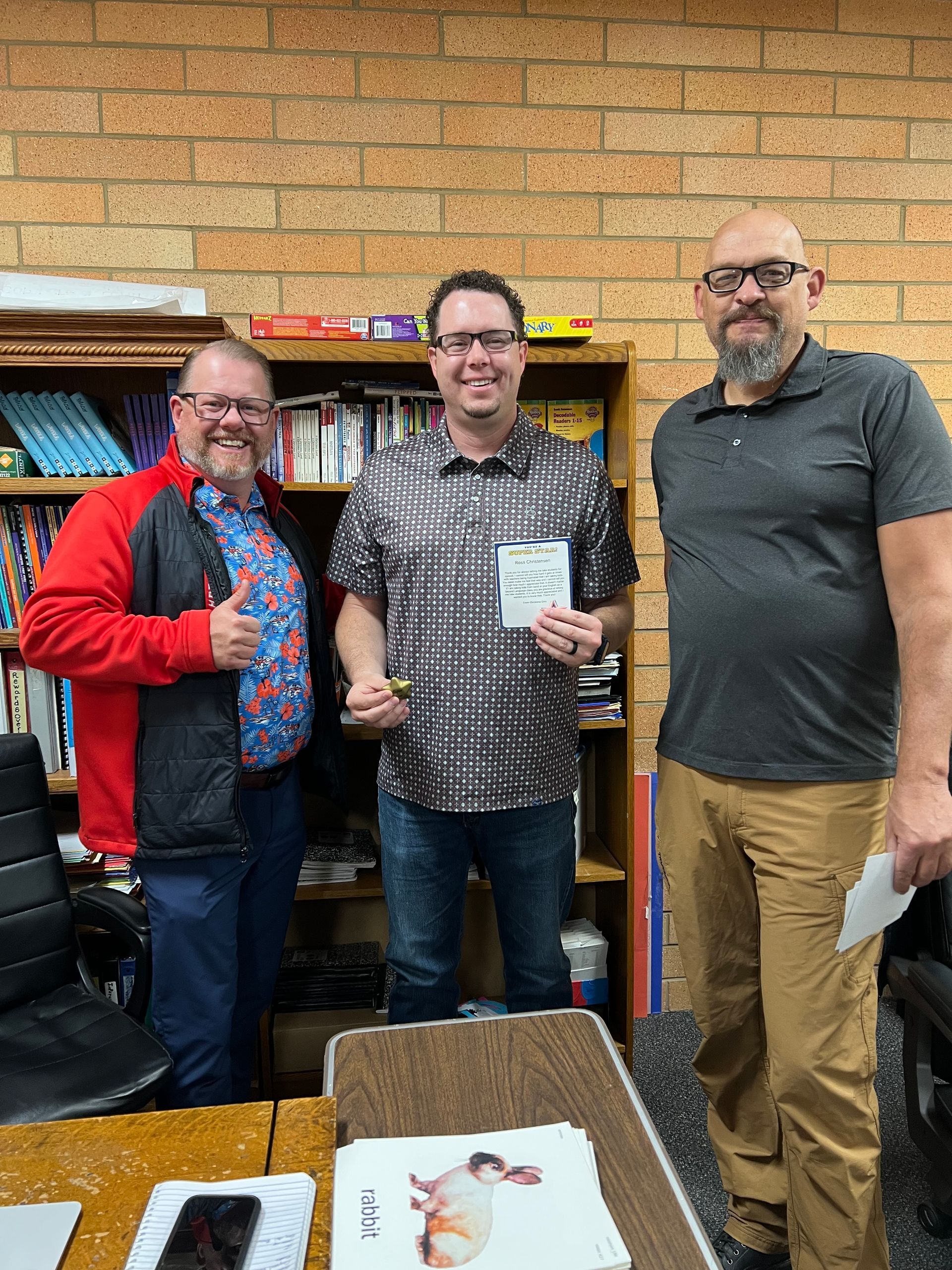 Three people standing in a library, smiling and holding papers in front of bookshelves.