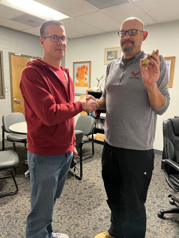 Two men shaking hands in an office. One in red sweater, the other in gray shirt holds a small object.