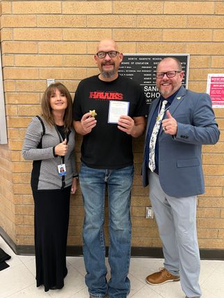 Three people stand in front of a brick wall. A man holds an award; another man gives a thumbs-up.