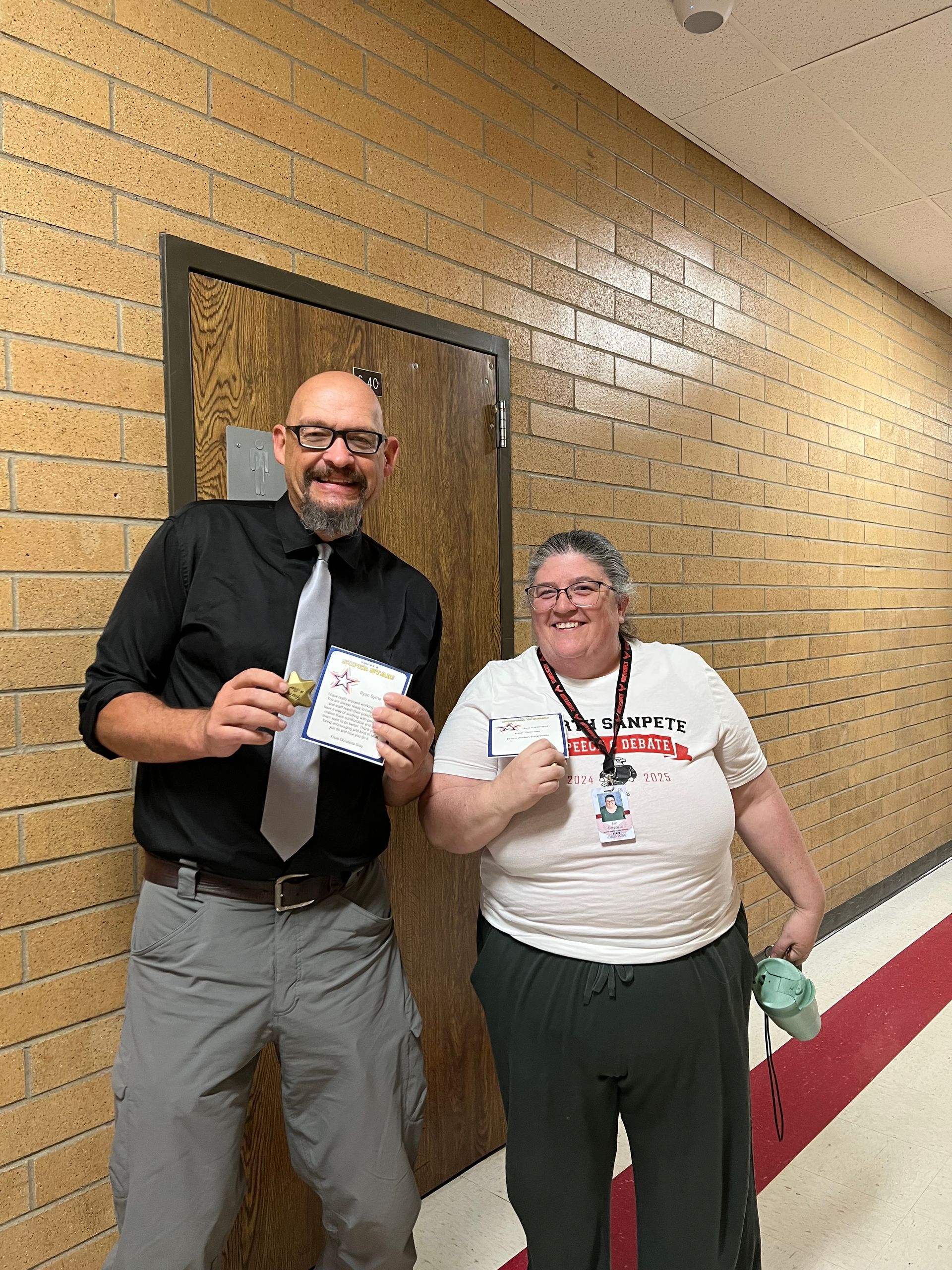 A man and a woman stand in a hallway, smiling and holding a medal and certificate.