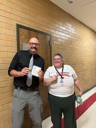 Man with tie holds award, smiles next to woman wearing lanyard, in hallway.