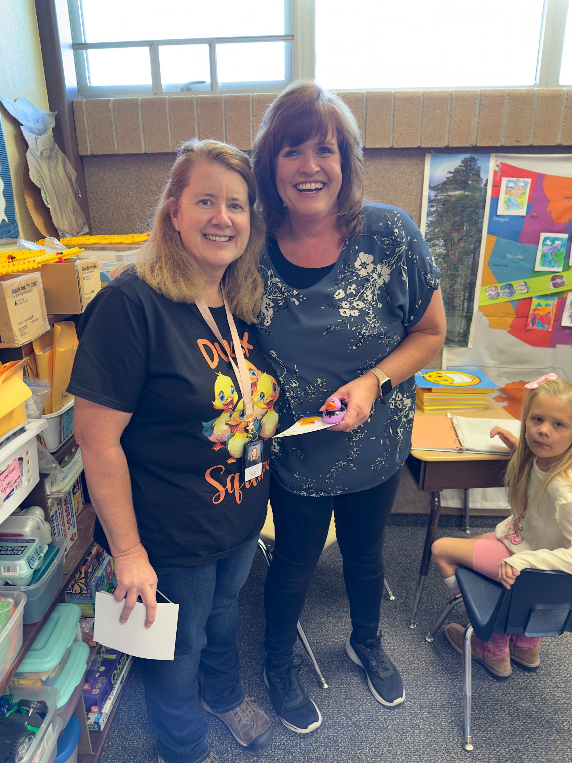 Two women are standing next to each other in a classroom and smiling