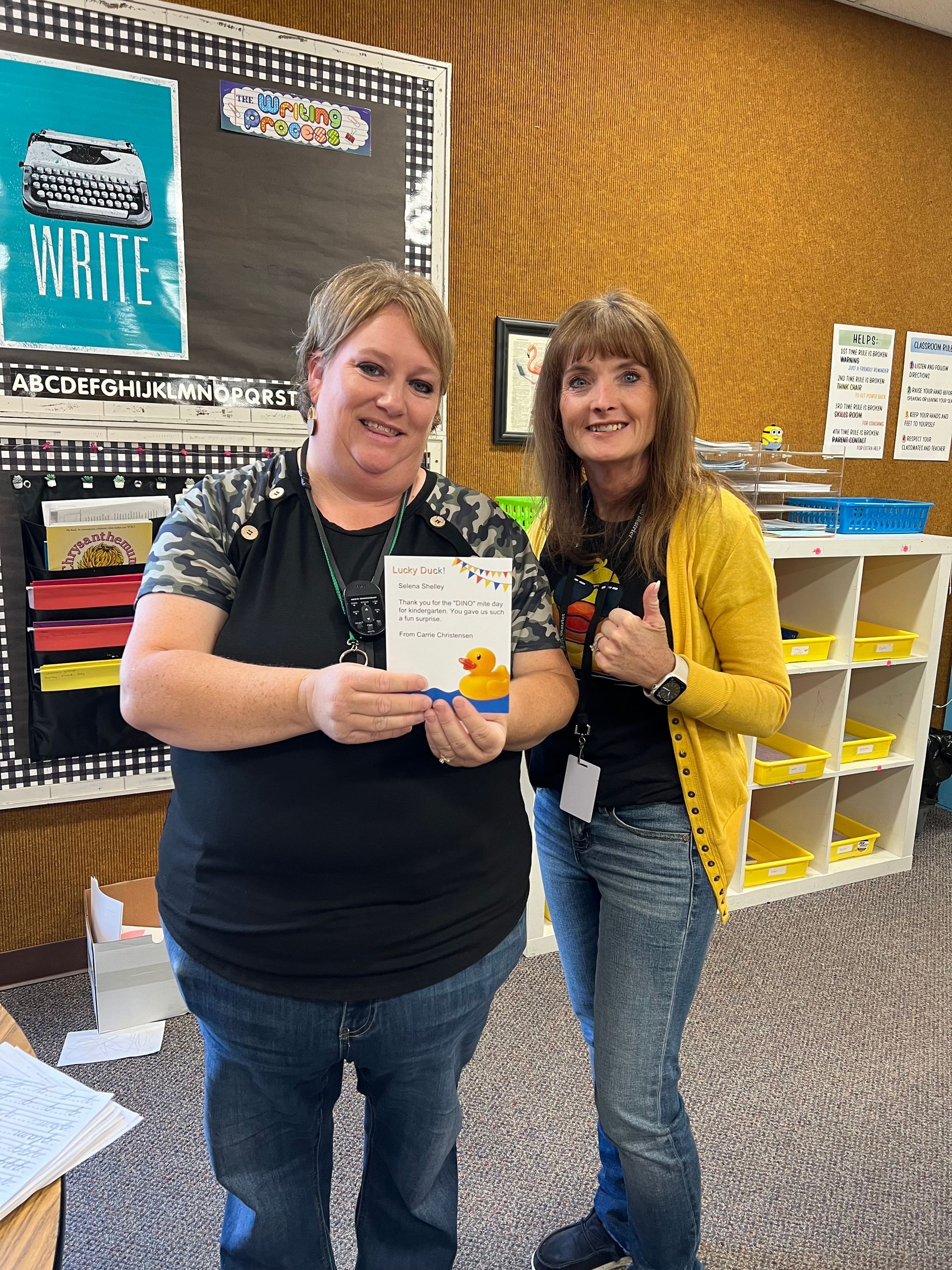 Two women are standing next to each other in a classroom holding a card.