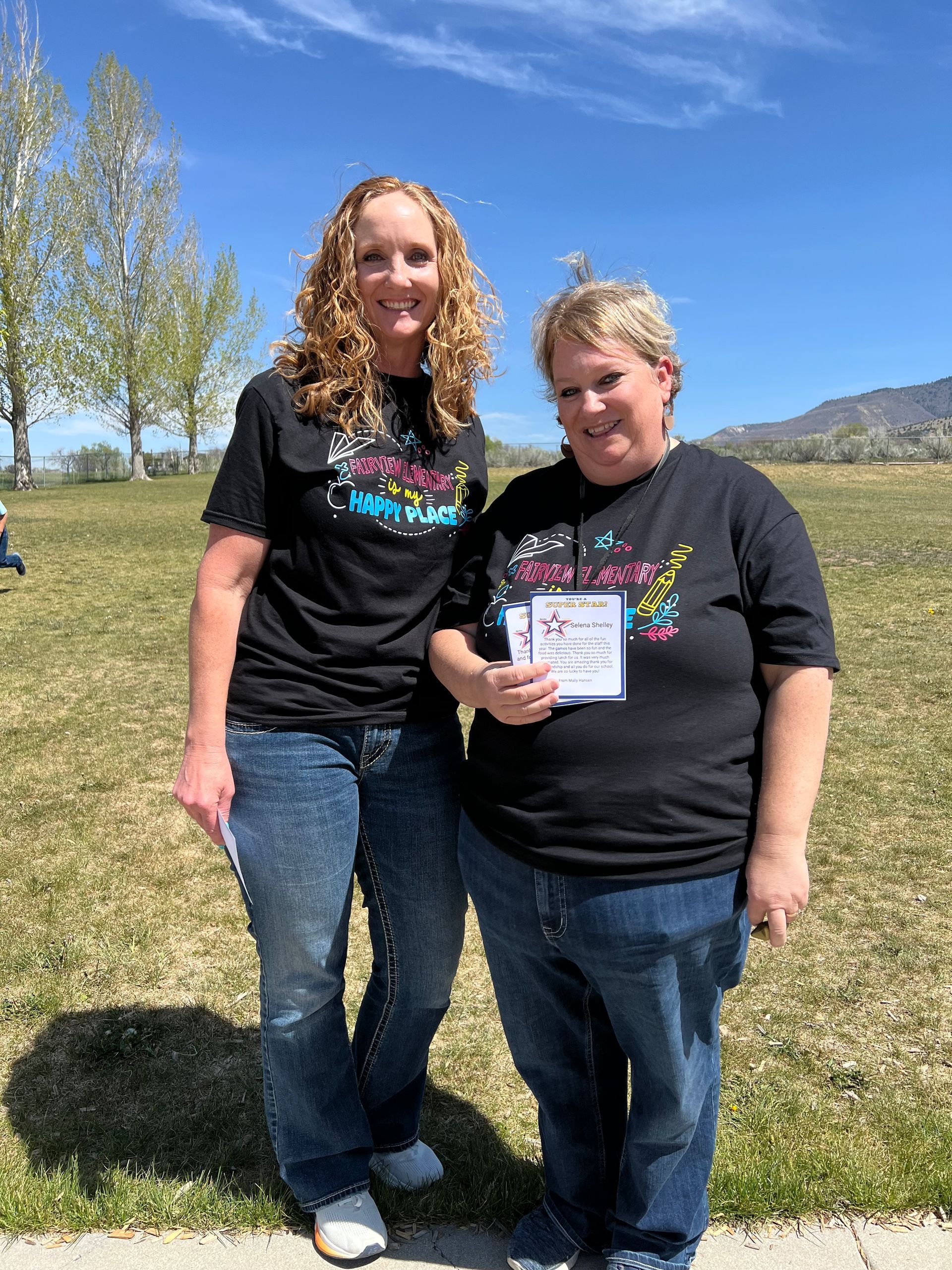 Two women in black T-shirts and jeans stand smiling outdoors, holding a sign in a sunny grassy field.