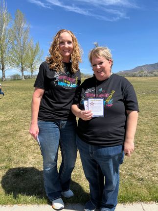 Two women in black T-shirts smiling outdoors, one holding a small sign on a grassy field under a blue sky.