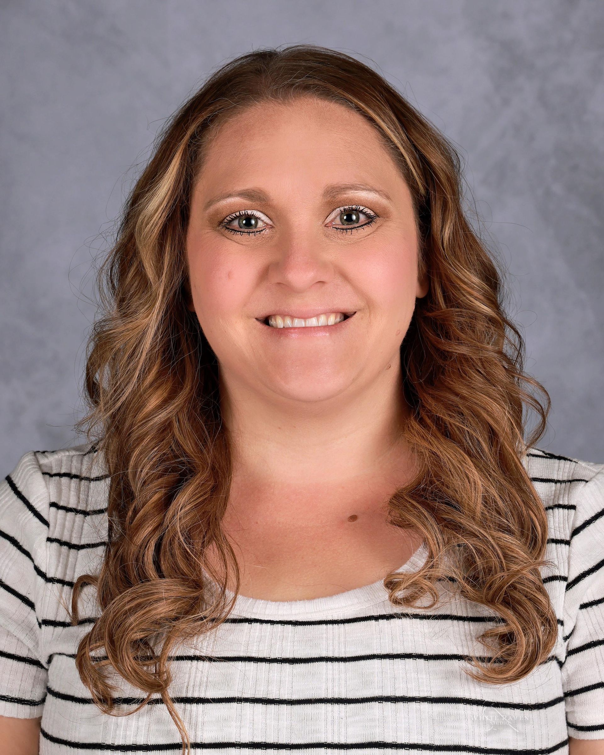 A smiling woman with wavy brown hair wearing a white shirt with black horizontal stripes against a grey backdrop.