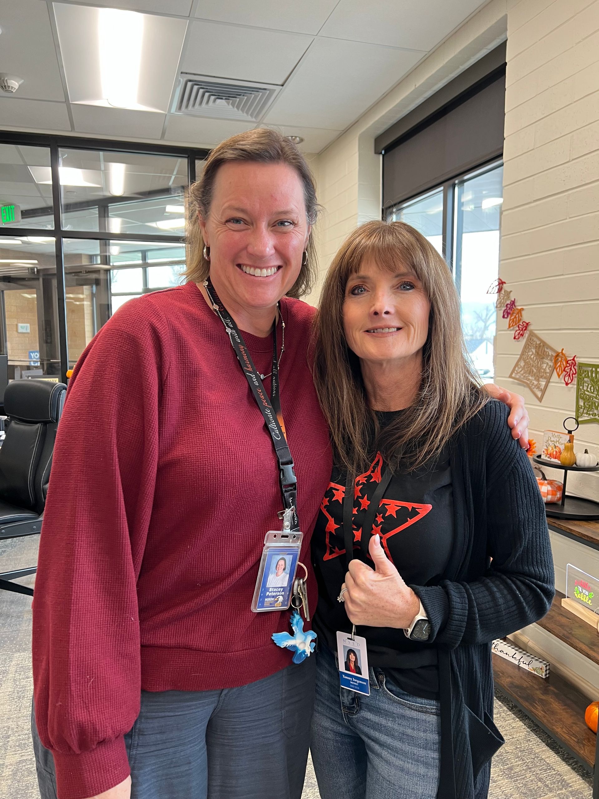 Two women smiling, inside a building. One wears red, the other a black shirt and cardigan. Both have lanyards.