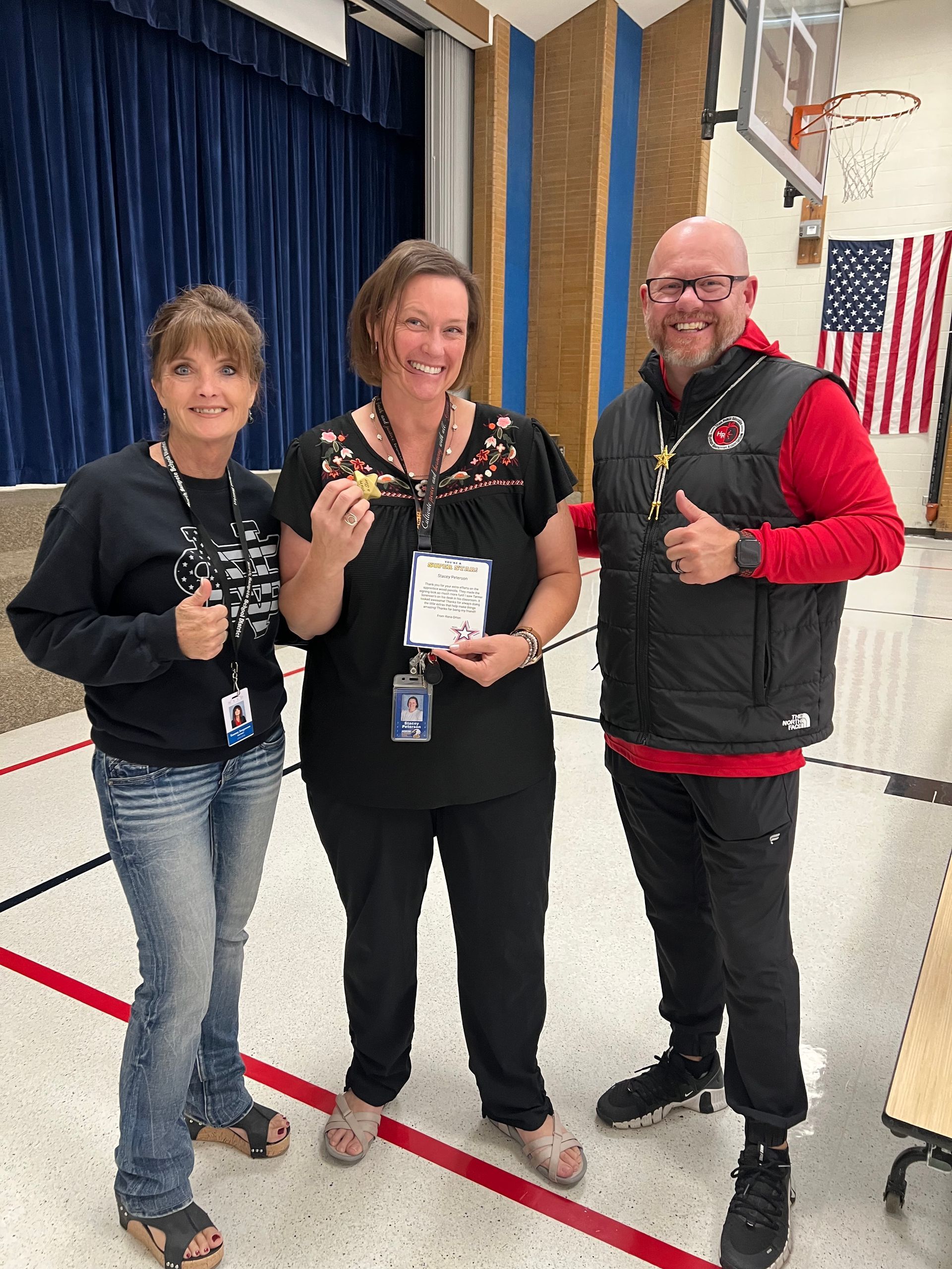 Three adults in a school gym, smiling and holding a key and award.