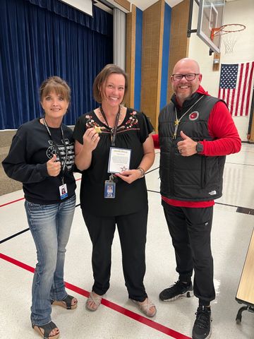 Three school staff smiling, holding a key and award, standing in a gymnasium.